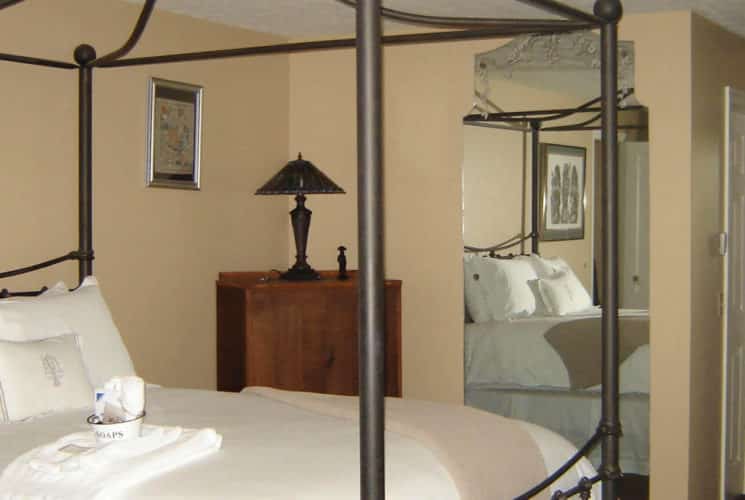 Graceful beige guestroom with a large iron canopy bed made up in white bedding, a dresser and mirror.